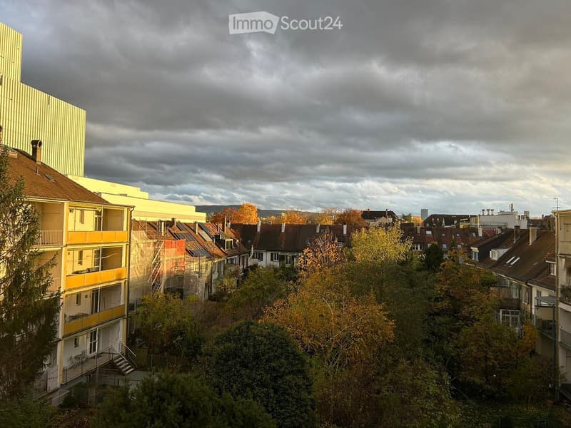 A multi-story residential building with yellow and white facades, surrounded by trees and neighboring houses with red-tiled roofs.