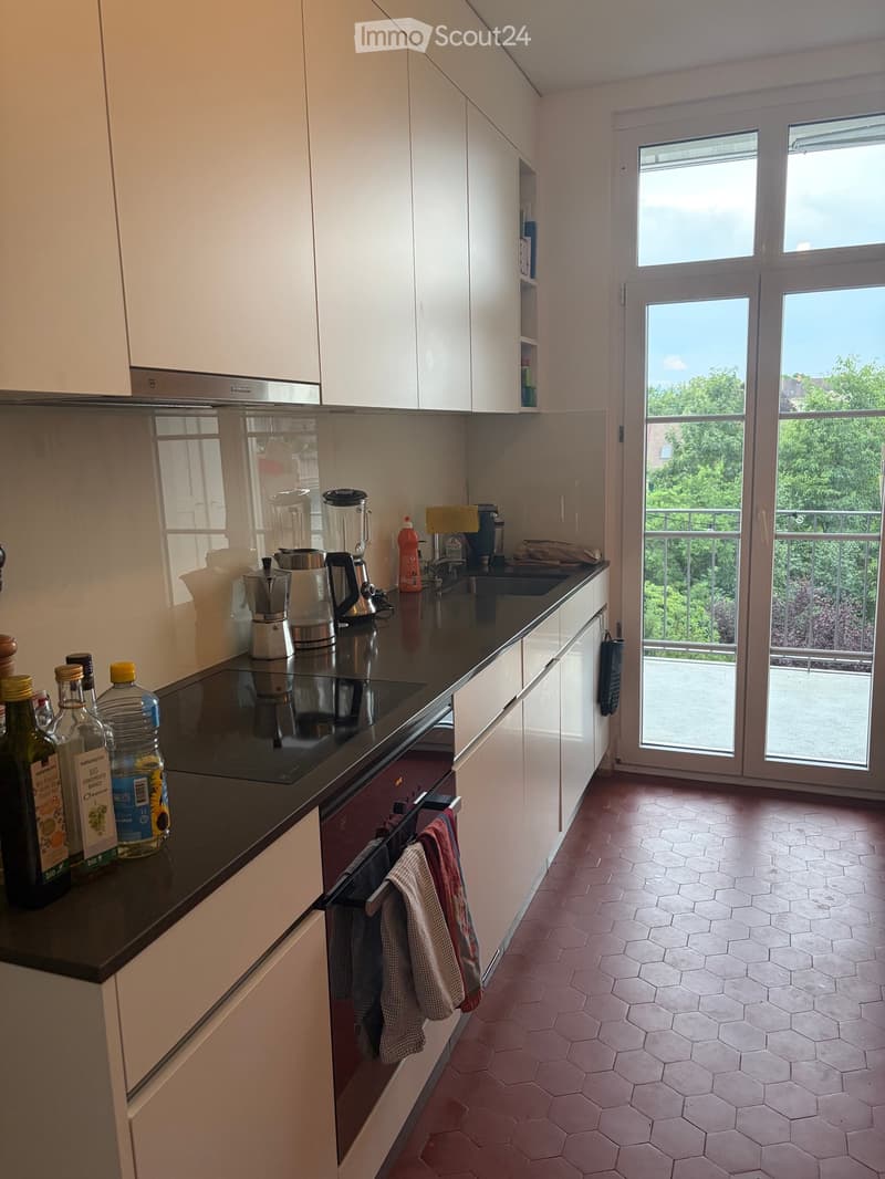 Modern kitchen with white cabinets, black countertop, induction stove, coffee maker, blender, and a view of a balcony through large windows.