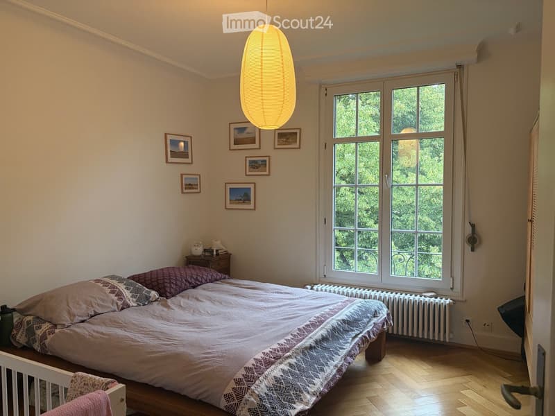 Cozy bedroom with a double bed, nightstand, radiator, and framed pictures on the wall. The room features wooden flooring and large windows with a view of greenery.