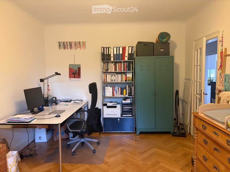 A well-organized office space with a wooden floor, a large desk with a computer and office supplies, a black office chair, a bookshelf filled with books and binders, a green cabinet, and a small wooden side table with a drawer.