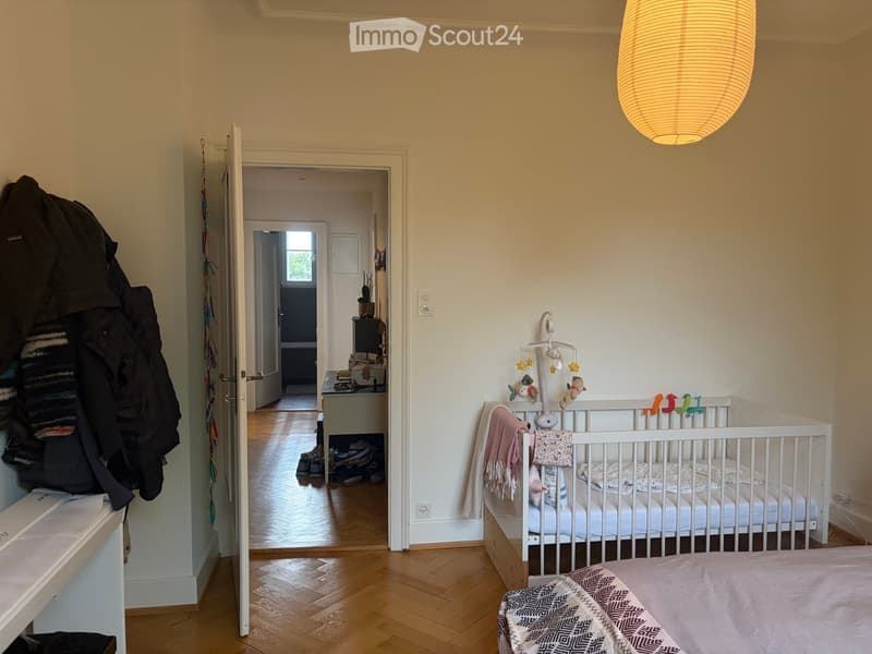 Brightly lit bedroom with a crib, toy mobile, and a bench with coats. The room has wooden flooring and an open doorway leading to another room.