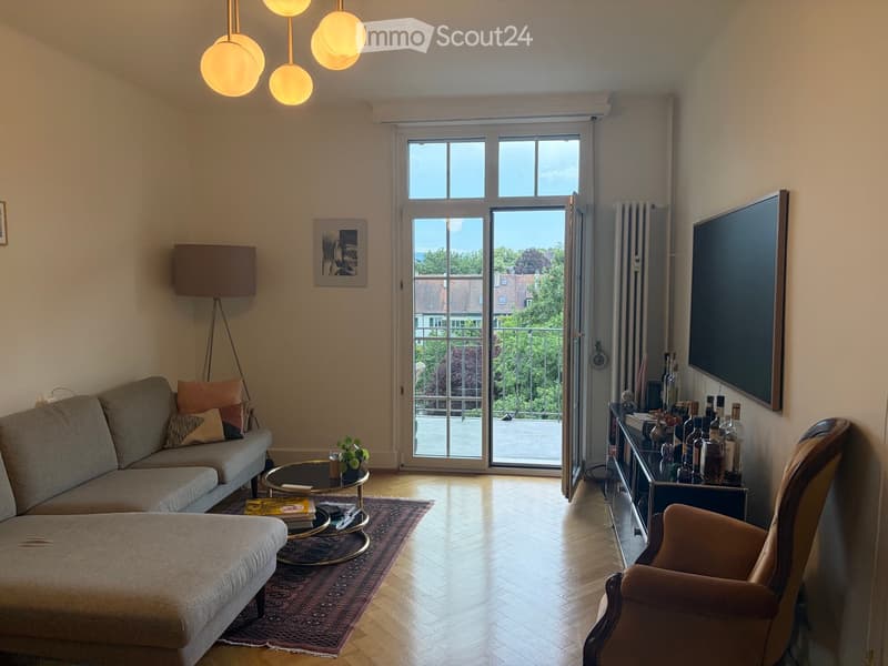 Modern living room with a grey sofa, ottoman, coffee table, armchair, and a TV. Features pendant lights, a floor lamp, and a large window with a view of a balcony.