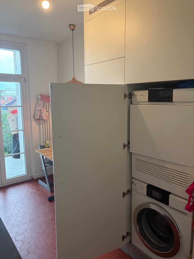 A small, modern room with a washing machine, a small table, and white cabinets. The room has a red tiled floor and a window with a view of the balcony.