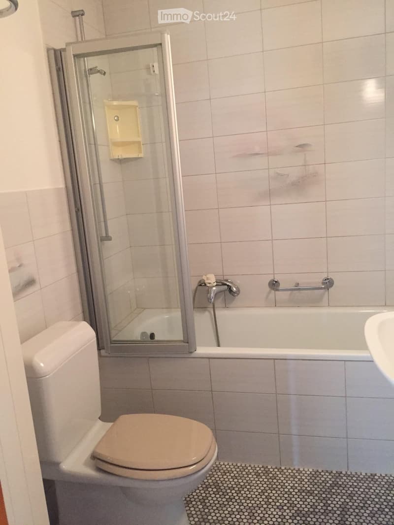 Bathroom with a toilet, bathtub, shower, and handrail. The floor has a patterned tile design, and the walls are tiled.