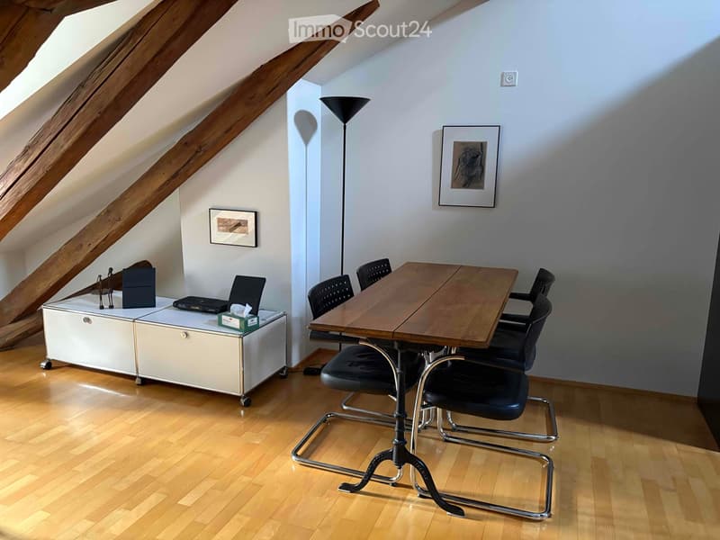 Bright attic room with wooden floors, slanted wooden ceiling beams, a large wooden table surrounded by black chairs, a white cabinet with a laptop, and two picture frames on the walls.