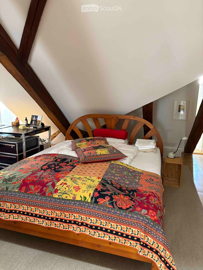 Cozy attic bedroom with wooden bed, colorful patchwork quilt, nightstand, small table, and framed picture on wall.