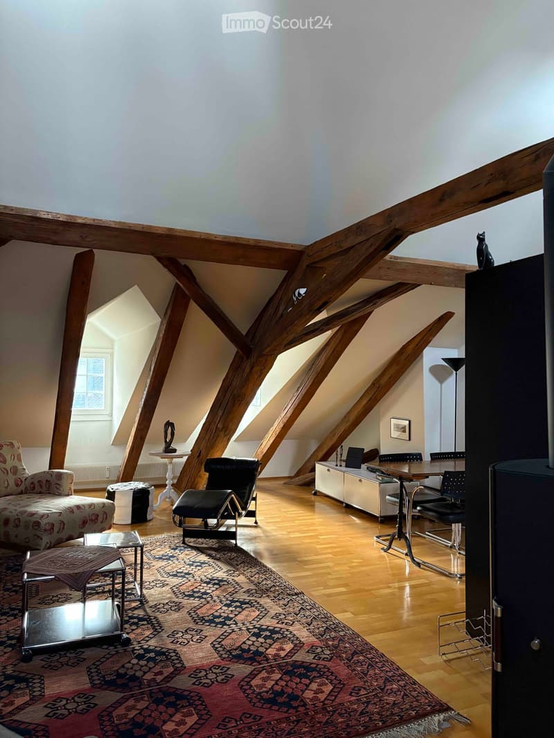 Spacious attic room with wooden beams, hardwood floors, and large windows. Features a patterned rug, a sofa, a leather chair, a small table with a sculpture, a cabinet with a laptop, and a dining area with chairs.