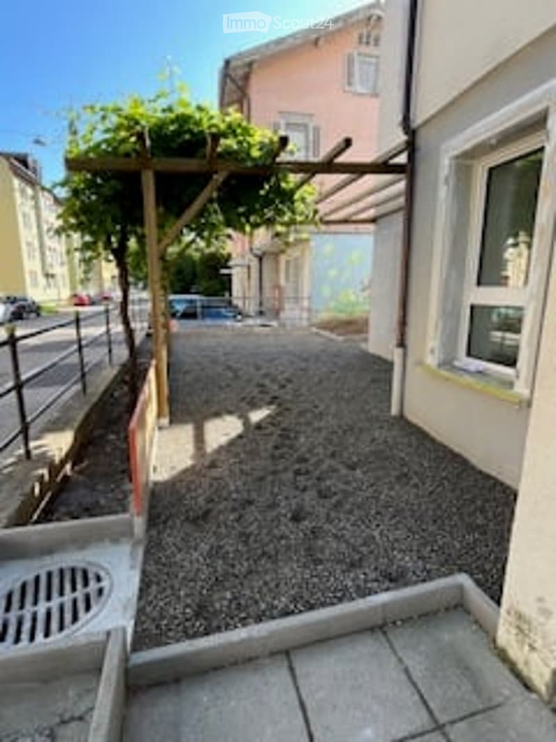 Small outdoor patio area with gravel ground, wooden pergola, and a few plants. Adjacent to a building with white walls and windows.