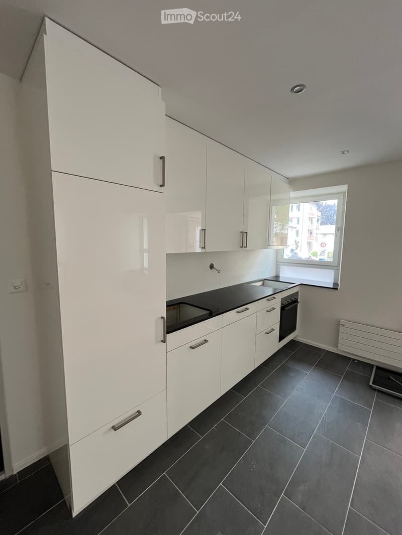 Modern kitchen with white cabinets, black countertop, and dark tiled floor. Features an oven and sink, with a window offering a view of the street.