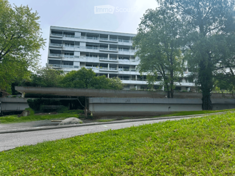 Multi-story residential building with multiple balconies and windows, surrounded by greenery and a paved pathway.