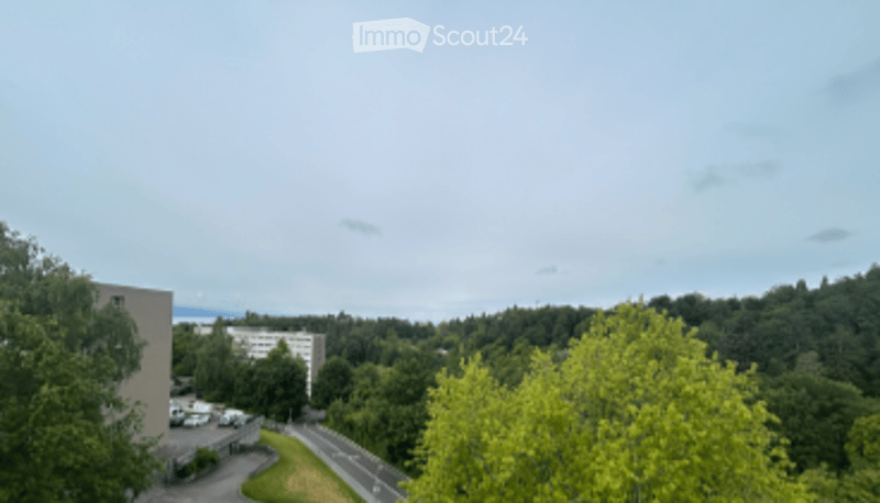 View from a balcony or terrace showing a road, parking area, trees, and distant buildings under a partly cloudy sky.