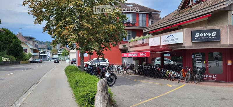 A commercial building with a storefront labeled "S+V+C" and "Ski + Velo-Center" with bicycles parked outside, surrounded by greenery and other buildings in the background.