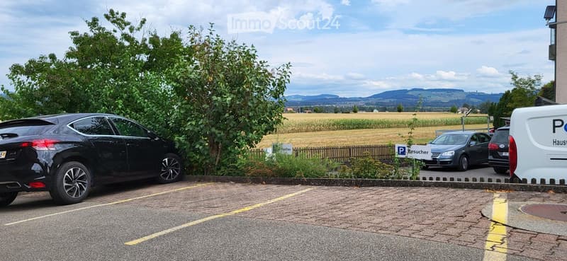Parking lot with several cars, surrounded by greenery and a rural landscape with fields and hills in the background.