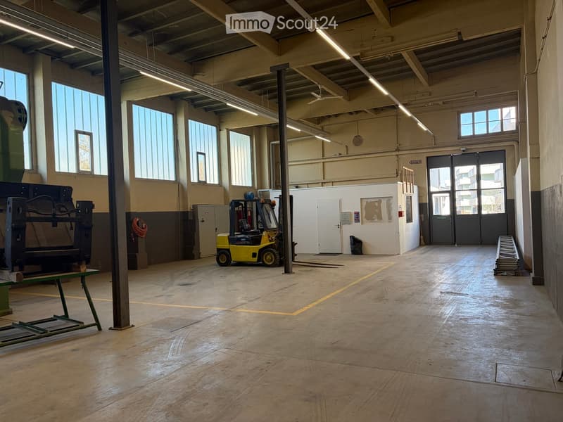 Large, empty industrial-style room with wooden flooring, large windows, and exposed metal beams. Contains a forklift, a green table, and a white partition wall.