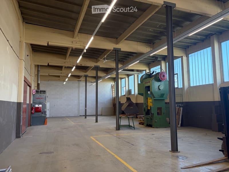 Large industrial workshop with high ceilings, exposed beams, and large windows. Contains a green industrial machine, a small table, a ladder, and a fire extinguisher.