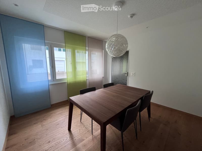 Dining area with wooden table, chairs, and a hanging lamp. Blue and green curtains on the left side.