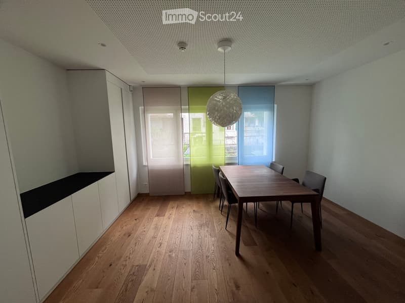 Dining area with wooden table, chairs, and a cabinet. White walls and wooden floor.