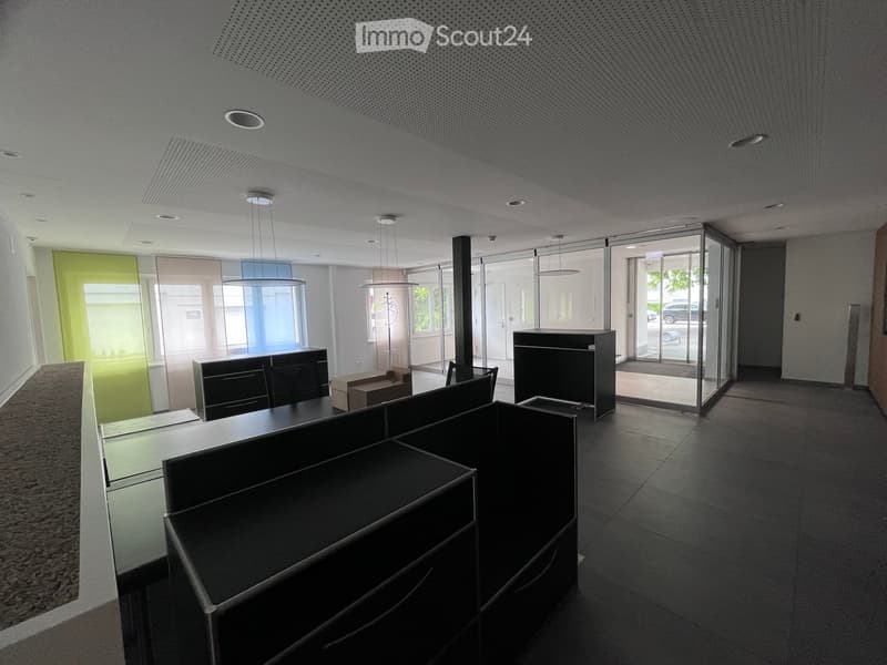 An empty room with black office cubicles, a black pillar, and a glass door leading to a parking area. The room has a white ceiling with a grid pattern and a carpeted floor.