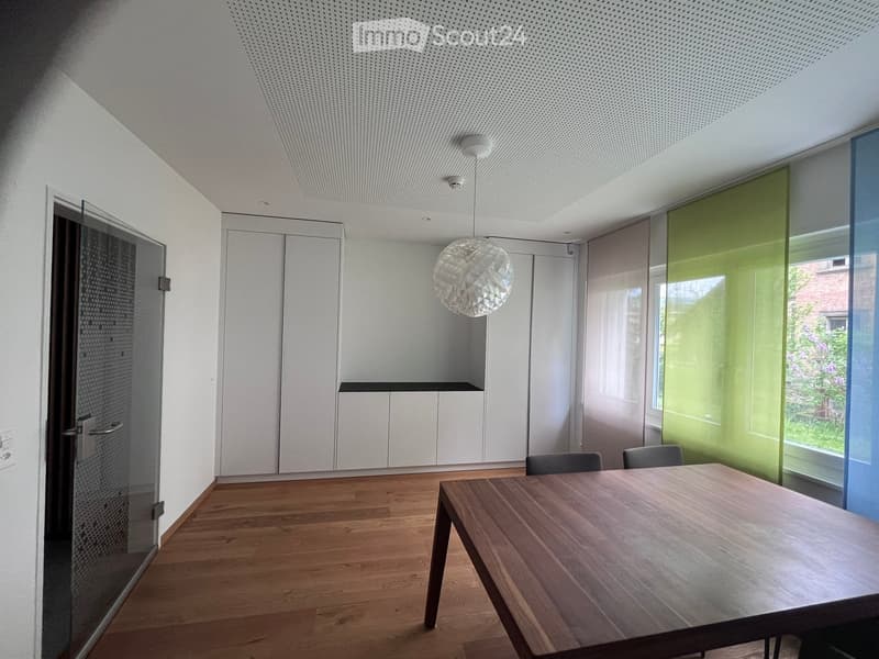 Modern, minimalist room with wooden flooring, a large wooden table, two chairs, white cabinets, and a decorative pendant light.