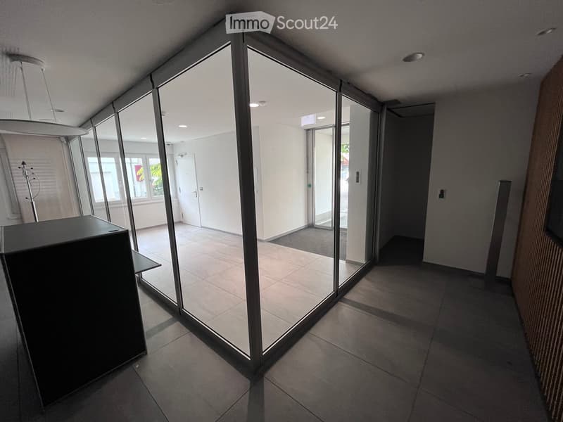 Modern hallway with large glass partitions, tiled floor, and recessed lighting. Features a reception desk on the left and a doorway leading to another area.