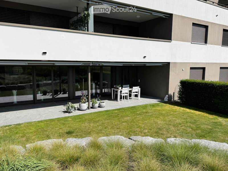 Modern house exterior with a well-maintained garden, featuring a patio area with a table and chairs, flower pots, and a neatly trimmed lawn.