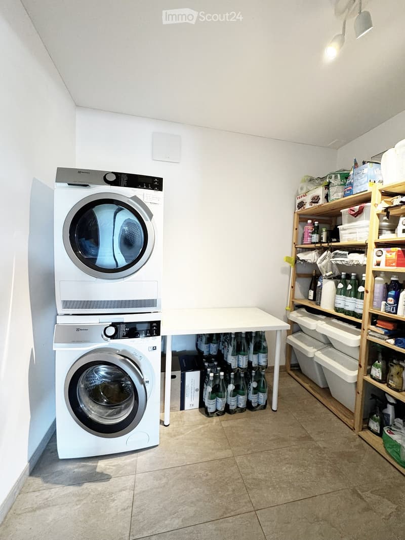 A small utility room with a stacked washing machine and dryer, a small table, and wooden shelves holding various items including bottled drinks and cleaning supplies.