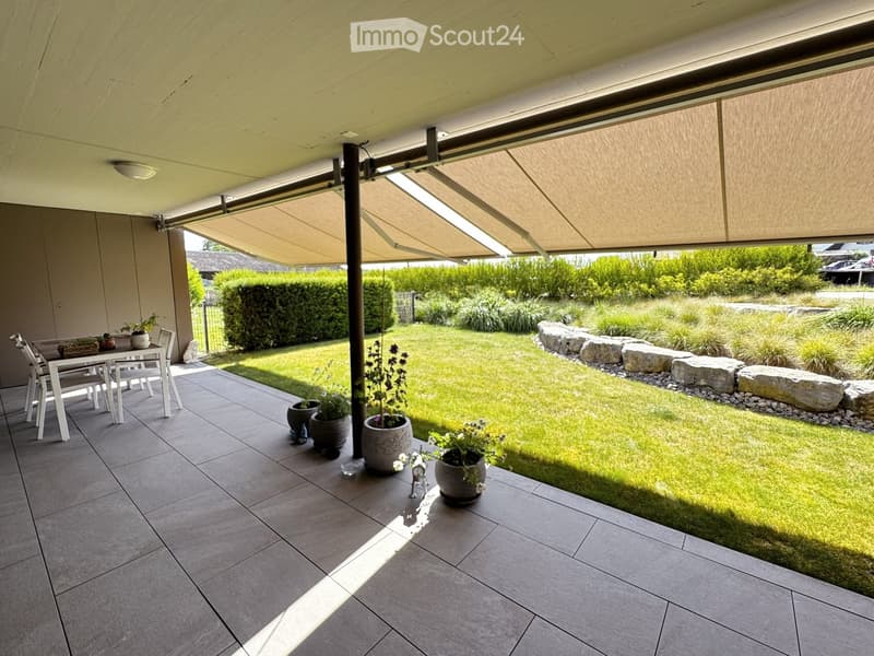 A covered patio area with a dining table and chairs, surrounded by a well-maintained garden with grass, shrubs, and decorative rocks.