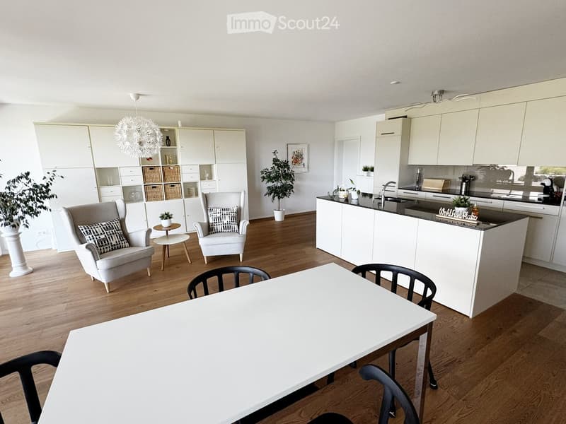 Open-plan living and dining area with a modern kitchen, white cabinets, a central island, and a dining table with chairs. The space includes a cozy seating area with armchairs and a small table.