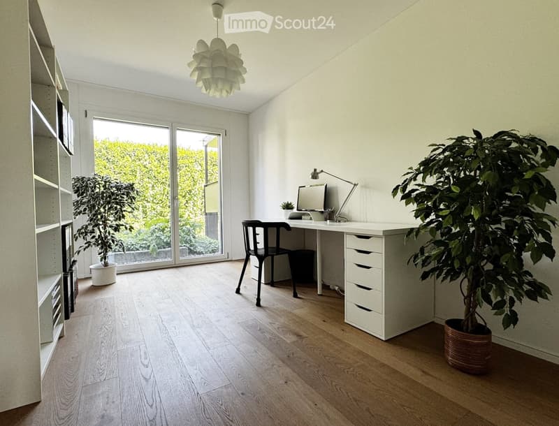 Bright, minimalist room with light wooden flooring, white walls, and large glass doors leading to a green outdoor area. Features a modern desk with a computer, a chair, a floor lamp, and two potted plants.