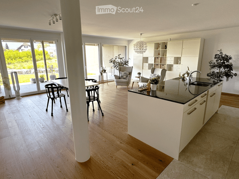 Modern kitchen and living area with light wood flooring, white cabinets, a central island, and large windows overlooking a green outdoor space.