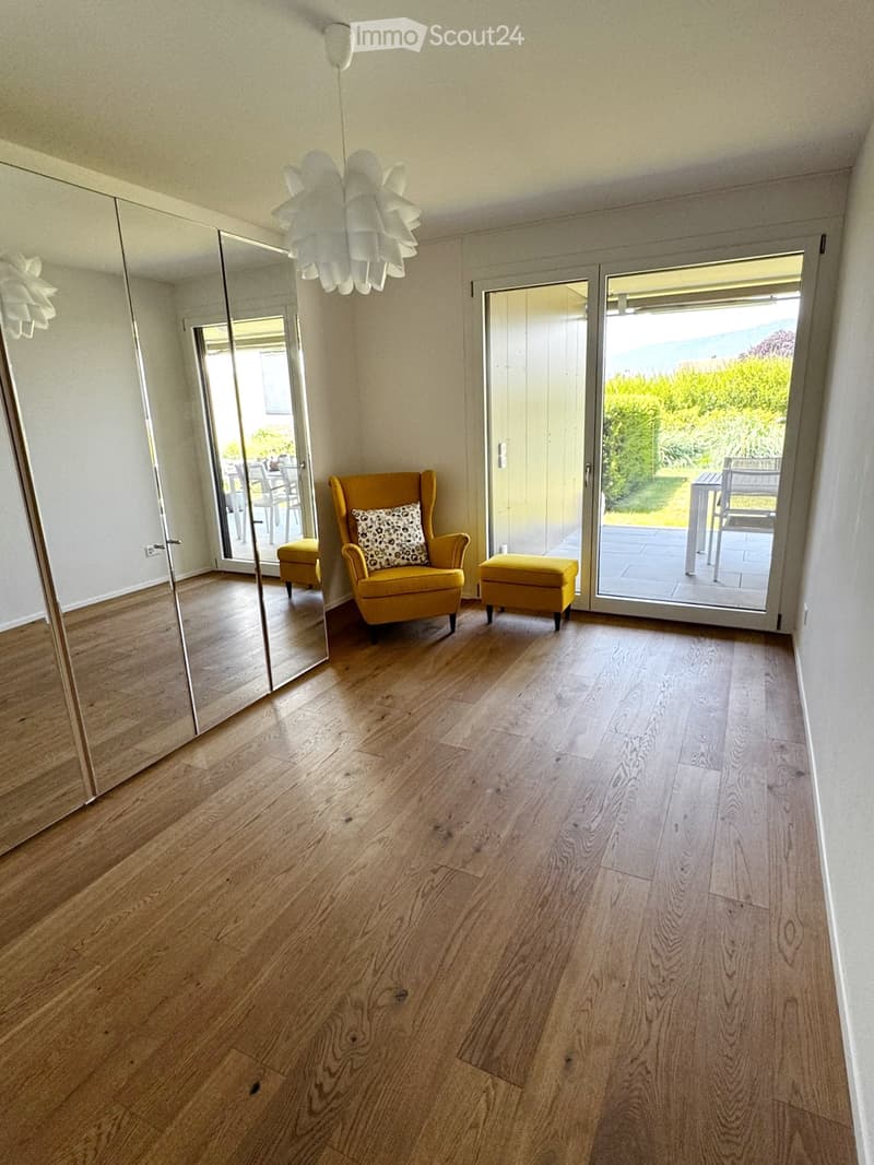 Bright living room with wooden flooring, large mirror, yellow armchair, matching stool, and pendant light fixture. Large sliding glass doors open to a balcony or terrace area.