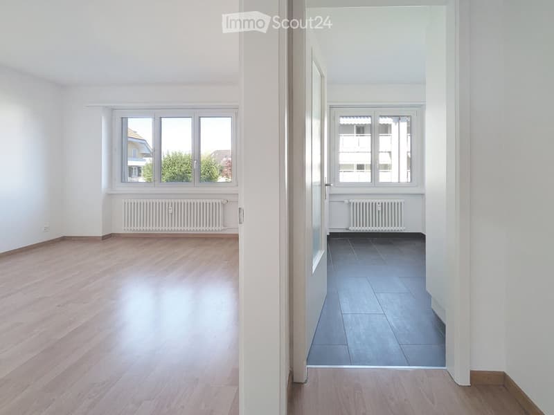 Empty unfurnished room with wooden flooring, white walls, and large windows. Another adjacent room with tiled flooring and white walls is visible through an open doorway.