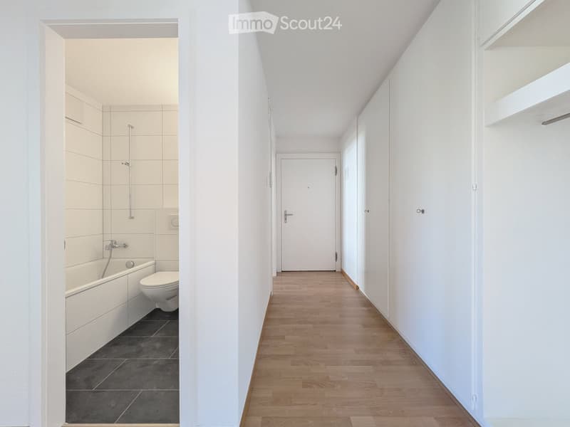 A narrow hallway with wooden flooring and white walls, leading to a bathroom with a bathtub, toilet, and tiled walls.