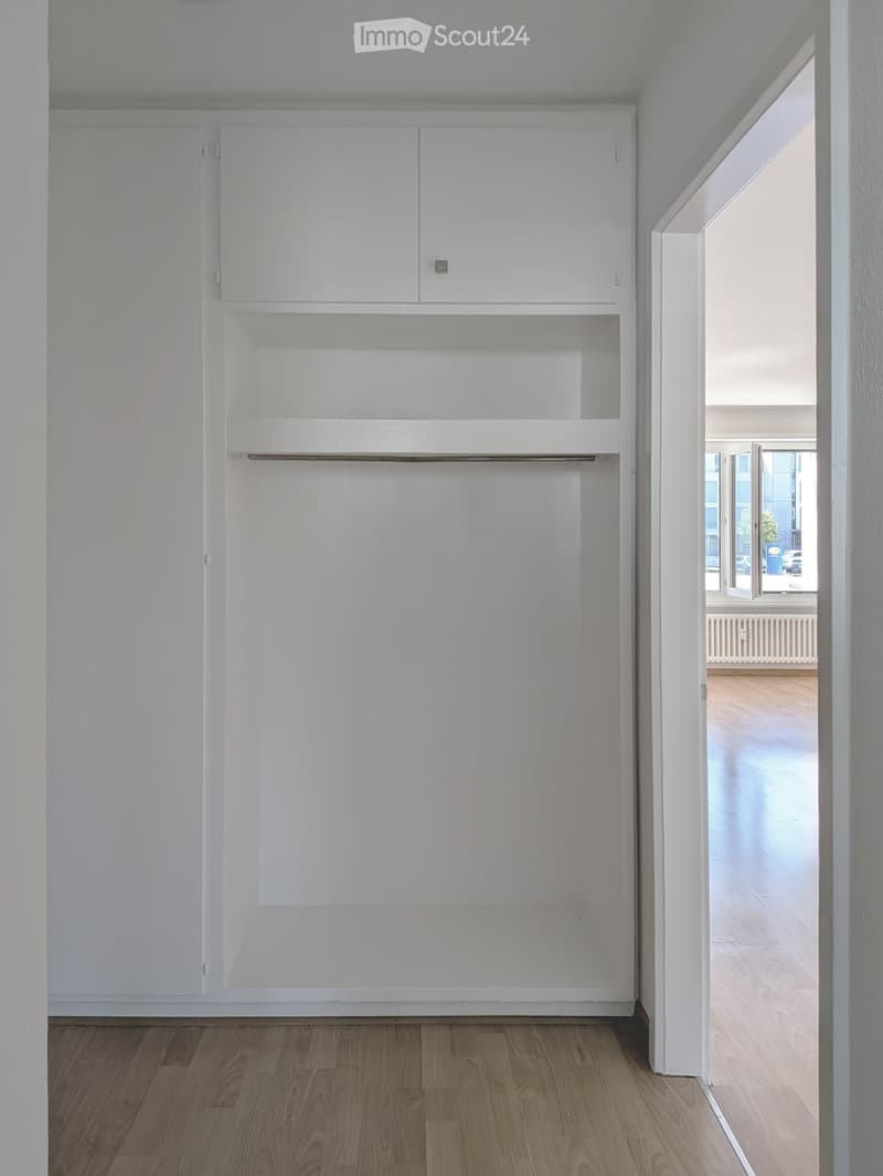 Empty room with white built-in cabinets and wooden flooring, leading to another room with large windows and wooden flooring.