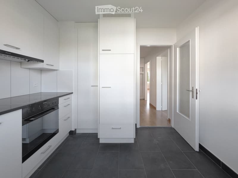 Modern hallway with white walls, dark tiled floor, and white cabinetry leading to a kitchen area with stainless steel appliances.