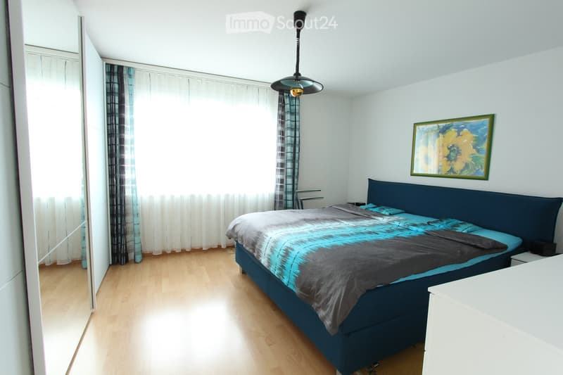 Modern bedroom with a large bed, nightstand, mirror, and painting on the wall. The room features wooden flooring and large windows with curtains.