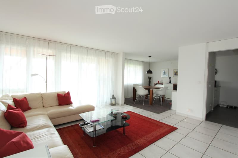 Bright living room with white walls, large windows with white curtains, a cream-colored sectional sofa with red cushions, a glass coffee table on a red rug, a floor lamp, and a dining area with a wooden table and white chairs.