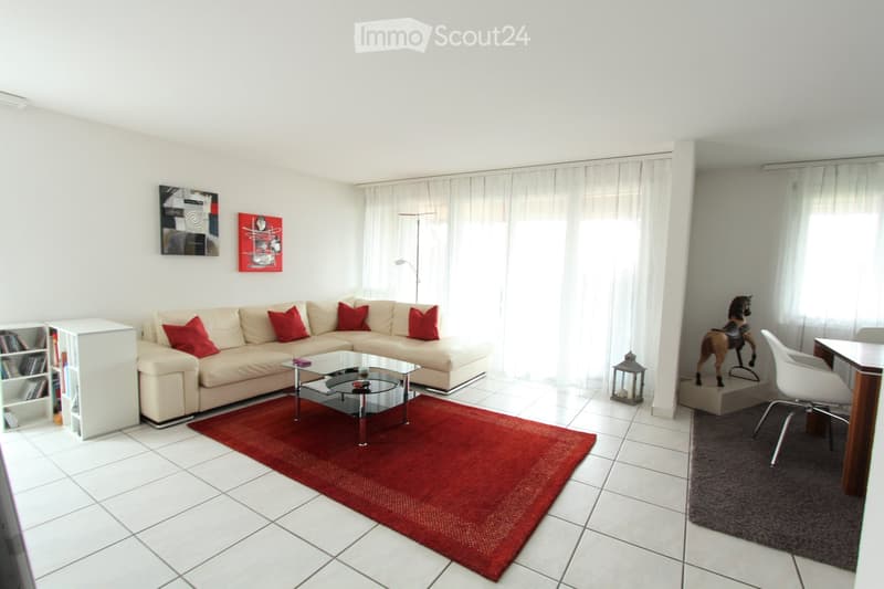 Bright living room with a beige sectional sofa, red throw pillows, a glass coffee table, a red area rug, a white bookshelf, a toy horse on a pedestal, and a small table with a white chair.