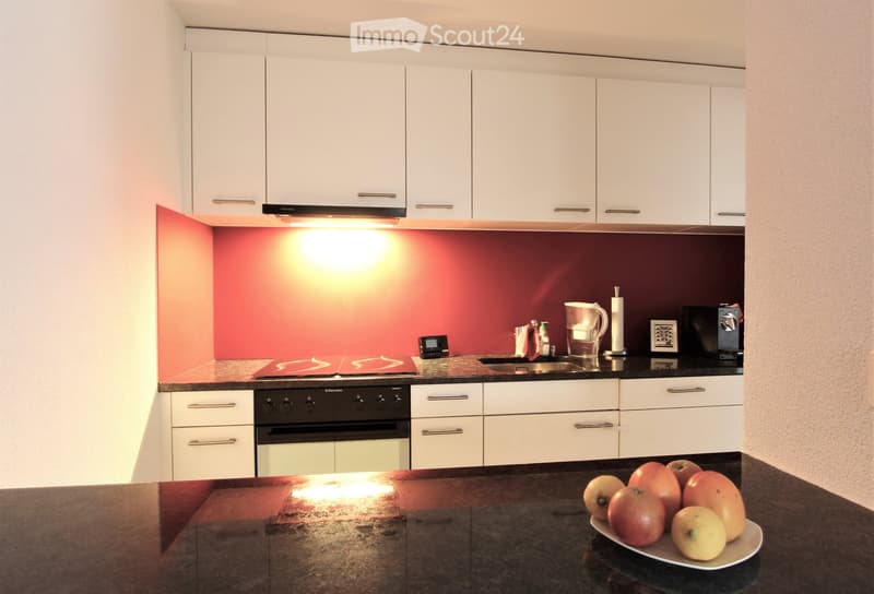 Modern kitchen with white cabinets, dark countertops, and a red accent wall. Features an oven, sink, faucet, water filter, coffee maker, and a plate of fruit on the counter.