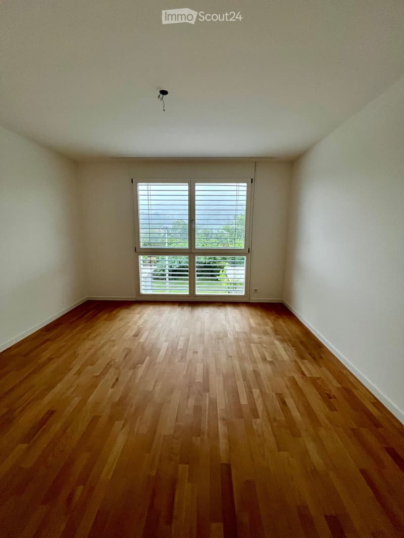 Empty room with wooden flooring and large windows with blinds, allowing natural light to enter.