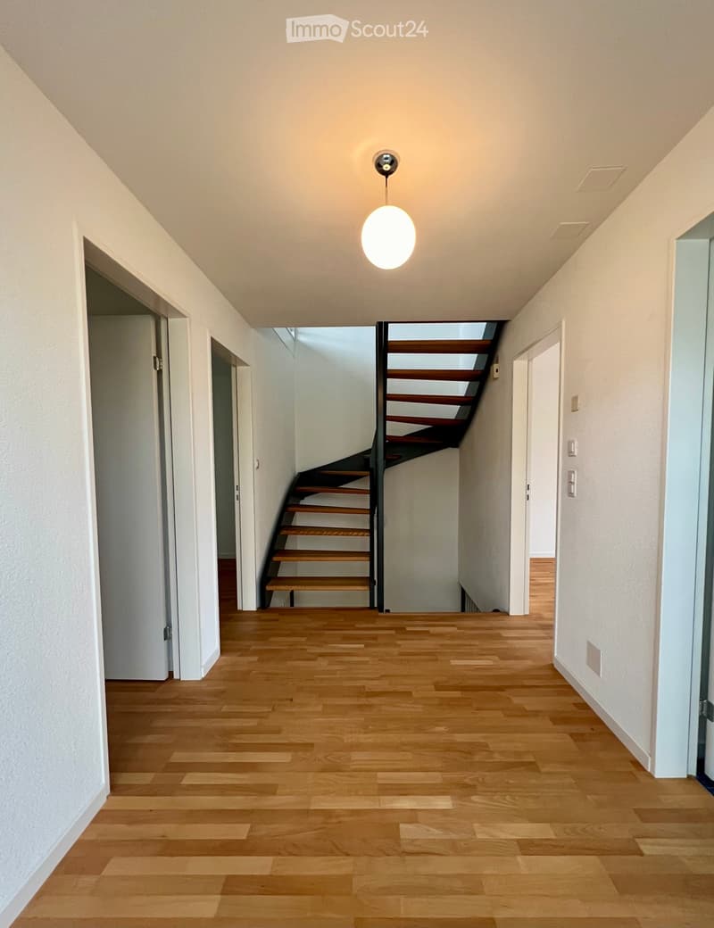 Bright hallway with wooden flooring, white walls, and a modern pendant light fixture. A staircase with wooden steps and a metal railing is visible at the end of the hallway.