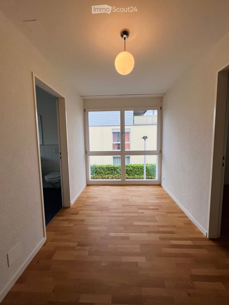 Empty hallway with wooden flooring, white walls, and a hanging light fixture. A door leads to a bathroom with a visible toilet, and a large window at the end offers a view of the outside.
