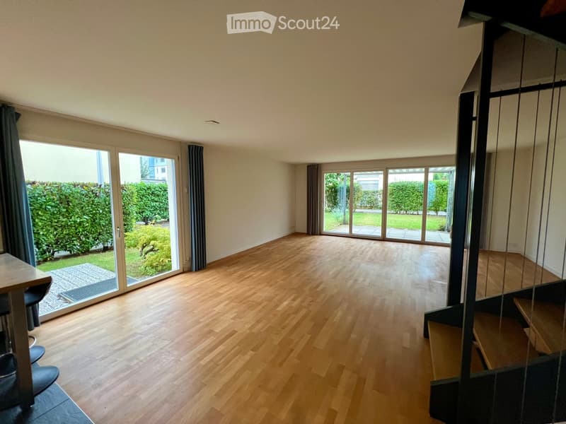 Bright, empty room with wooden flooring, large sliding glass doors leading to a garden, and a modern staircase visible on the right side.