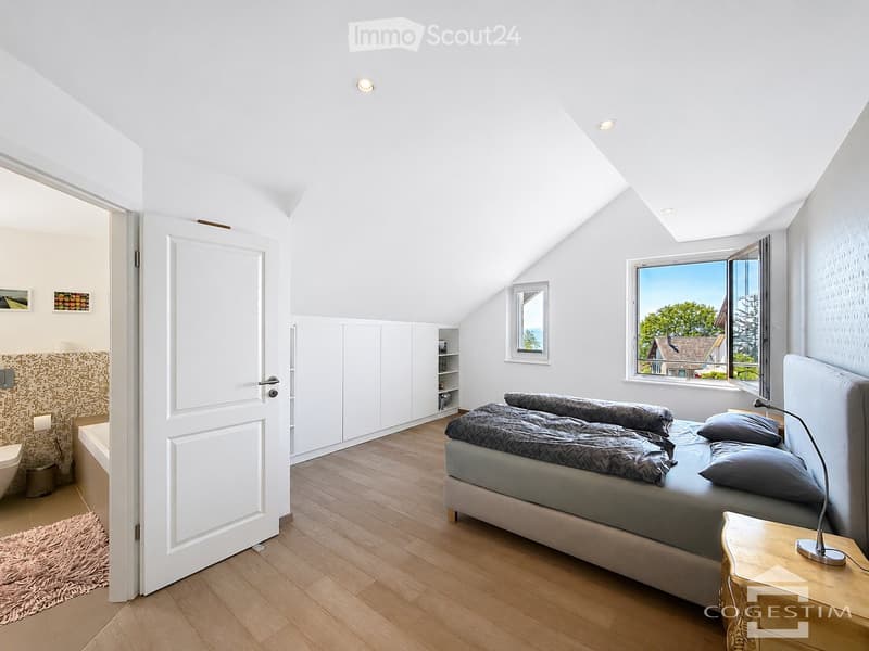 Bright, modern bedroom with wooden flooring, large window offering a view, bed with grey bedding, nightstand with lamp, built-in wardrobe, and an adjacent bathroom with a toilet and rug.
