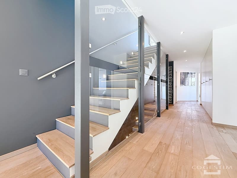 Corridor / Hallway. Modern hallway with wooden flooring, grey walls, and a staircase with glass railings leading to an upper level.
