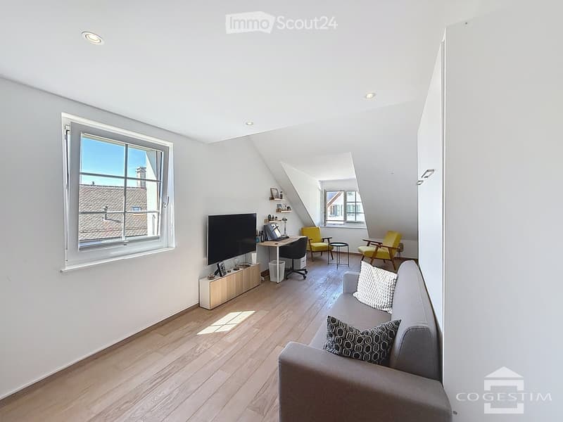 Furnished Room. Bright, modern living space with wooden flooring, large windows, a grey sofa, a yellow armchair, a small table, and a desk with a laptop and screen.