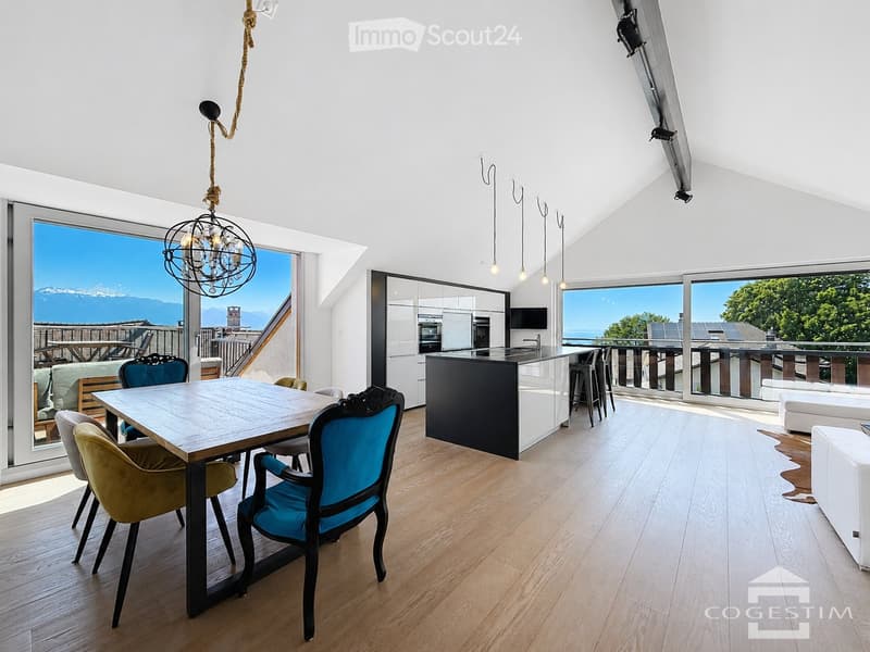 Open-plan living and dining area with wooden flooring, large windows offering a scenic view, a dining table with chairs, a modern kitchen island, and a chandelier.