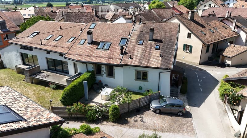A two-story house with a tiled roof, multiple windows, and a small garden area. A car is parked in the driveway, and the house is surrounded by neighboring buildings and greenery.