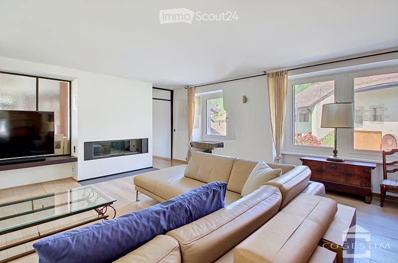 Modern living room with a beige leather sofa, glass coffee table, large flat-screen TV, fireplace, and large windows with curtains.