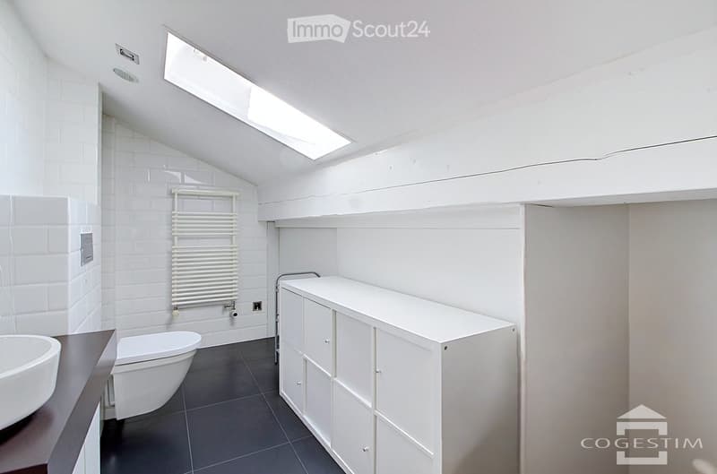 Modern bathroom with white tiles, a sink, toilet, built-in cabinet, and a radiator. The room features a skylight window and dark tiled flooring.